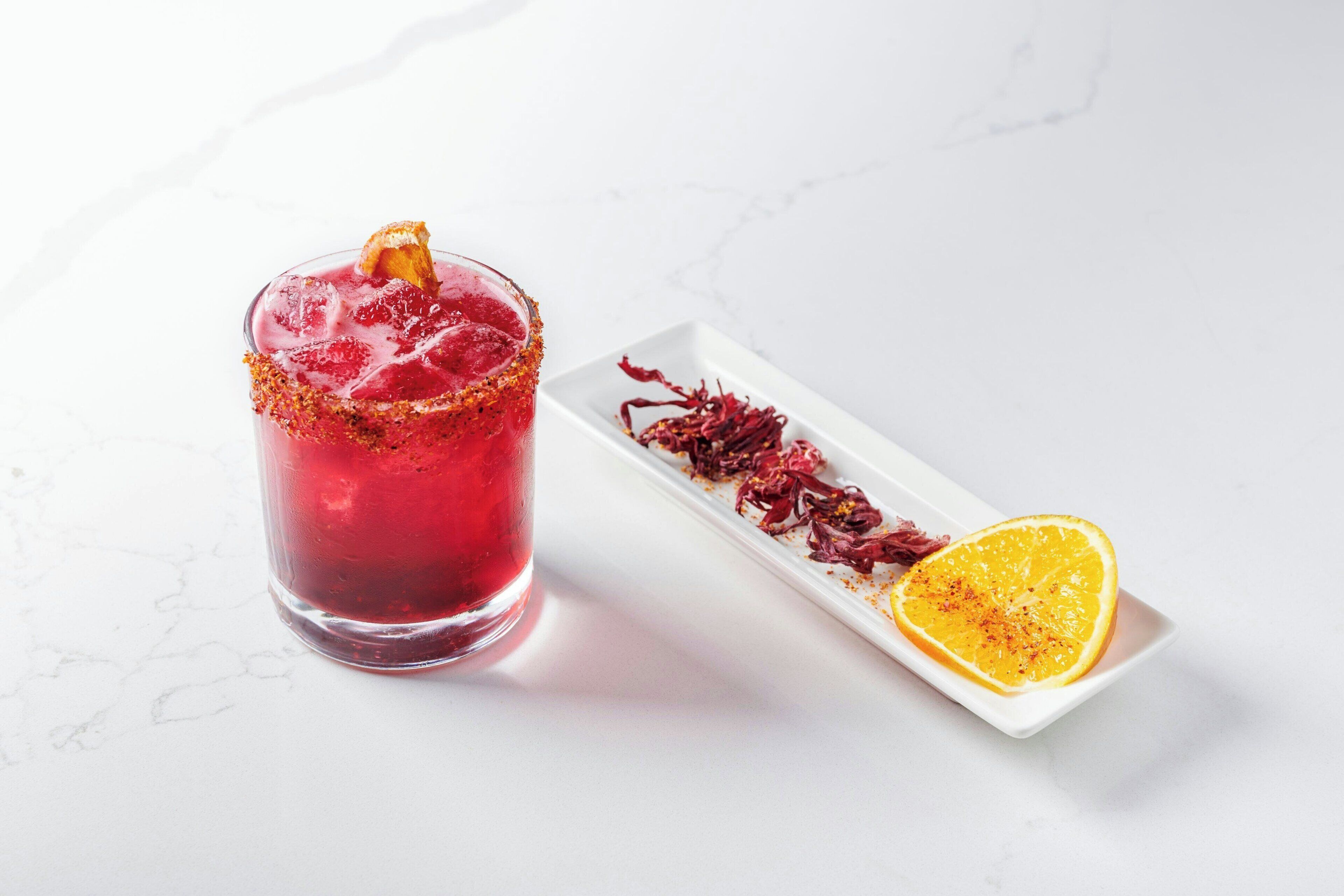 A glass of red drink with a garnish on a white plate beside it.