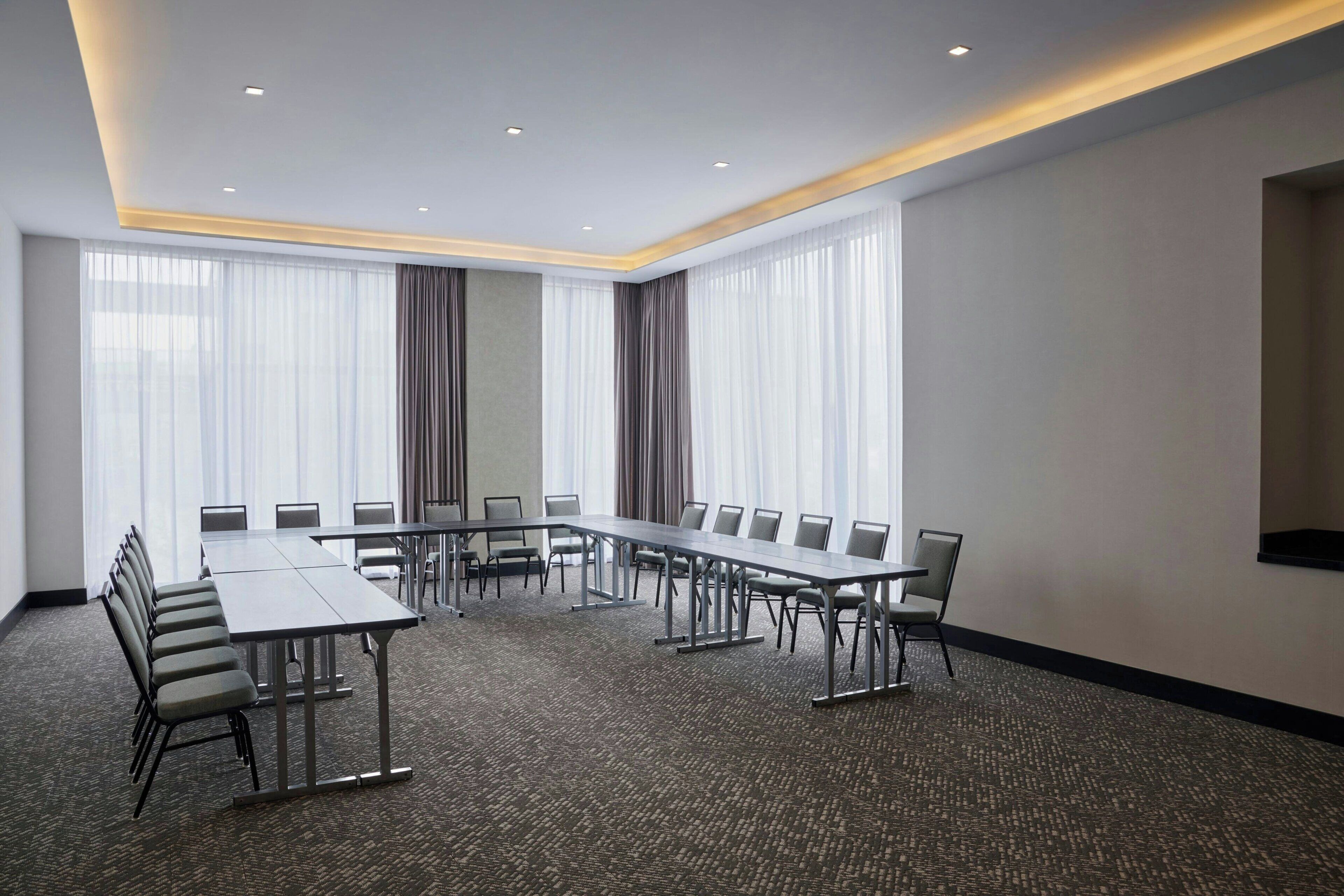A conference room with a long table and chairs, large windows with curtains, and a carpeted floor.