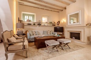 A cozy living room with a sofa, armchair, ottoman, and coffee table. There is a fireplace, a television, and shelves with books and decorative items.