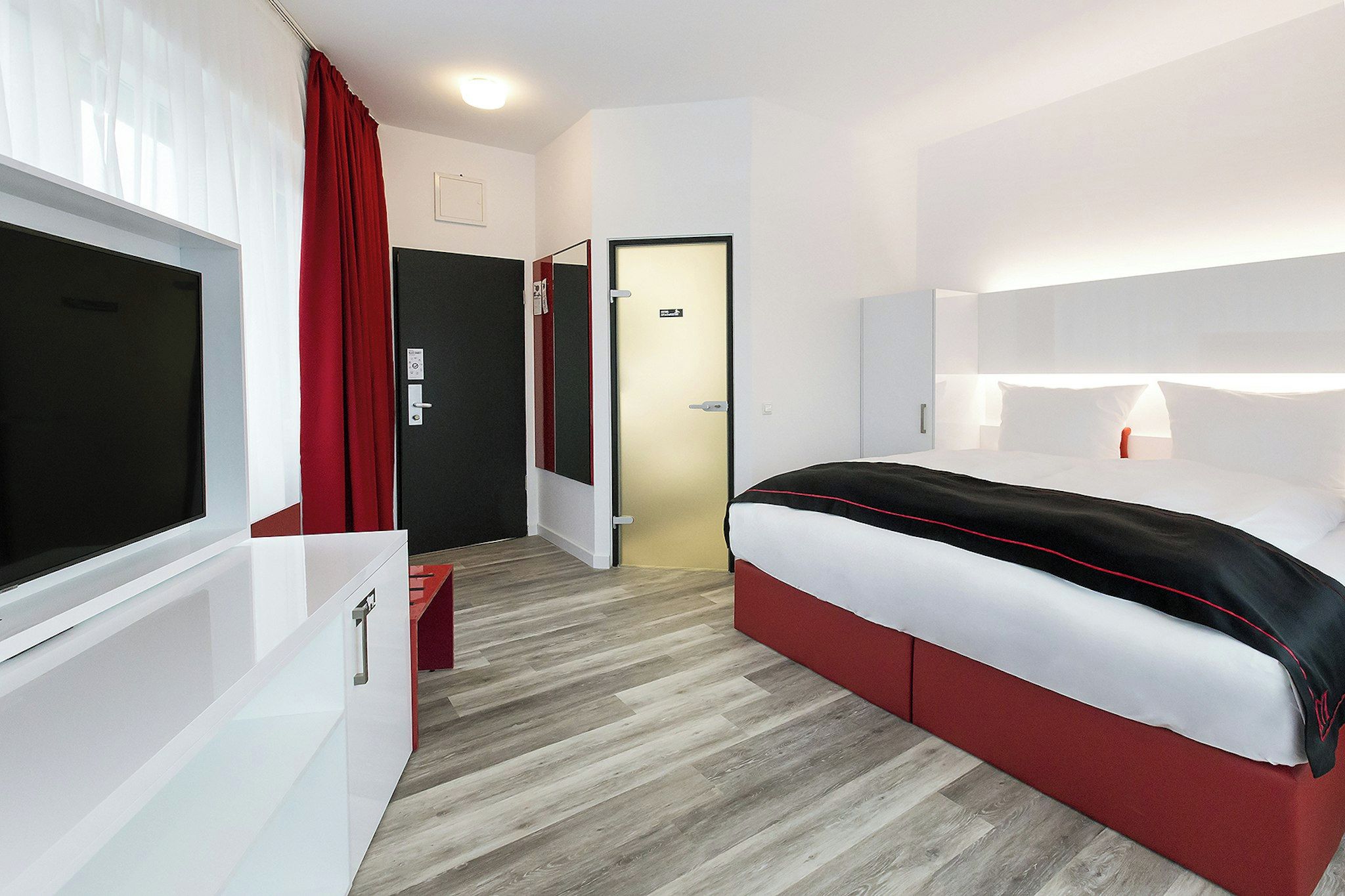 Ein modernes Hotelzimmer mit einem großen Bett, einem Fernseher, einem roten Vorhang und Holzboden.
