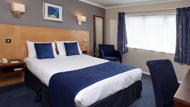 Une chambre d’hôtel avec un grand lit, un bureau, une chaise, une table de chevet et une lampe fixée au mur.