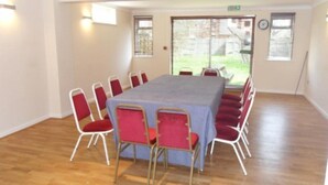 Une salle de conférence avec une longue table et des chaises rouges, de grandes fenêtres et une horloge au mur.