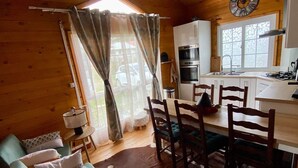 A cozy cabin interior with a wooden dining area, a kitchen with a clock, and a living room with a sofa and chairs.