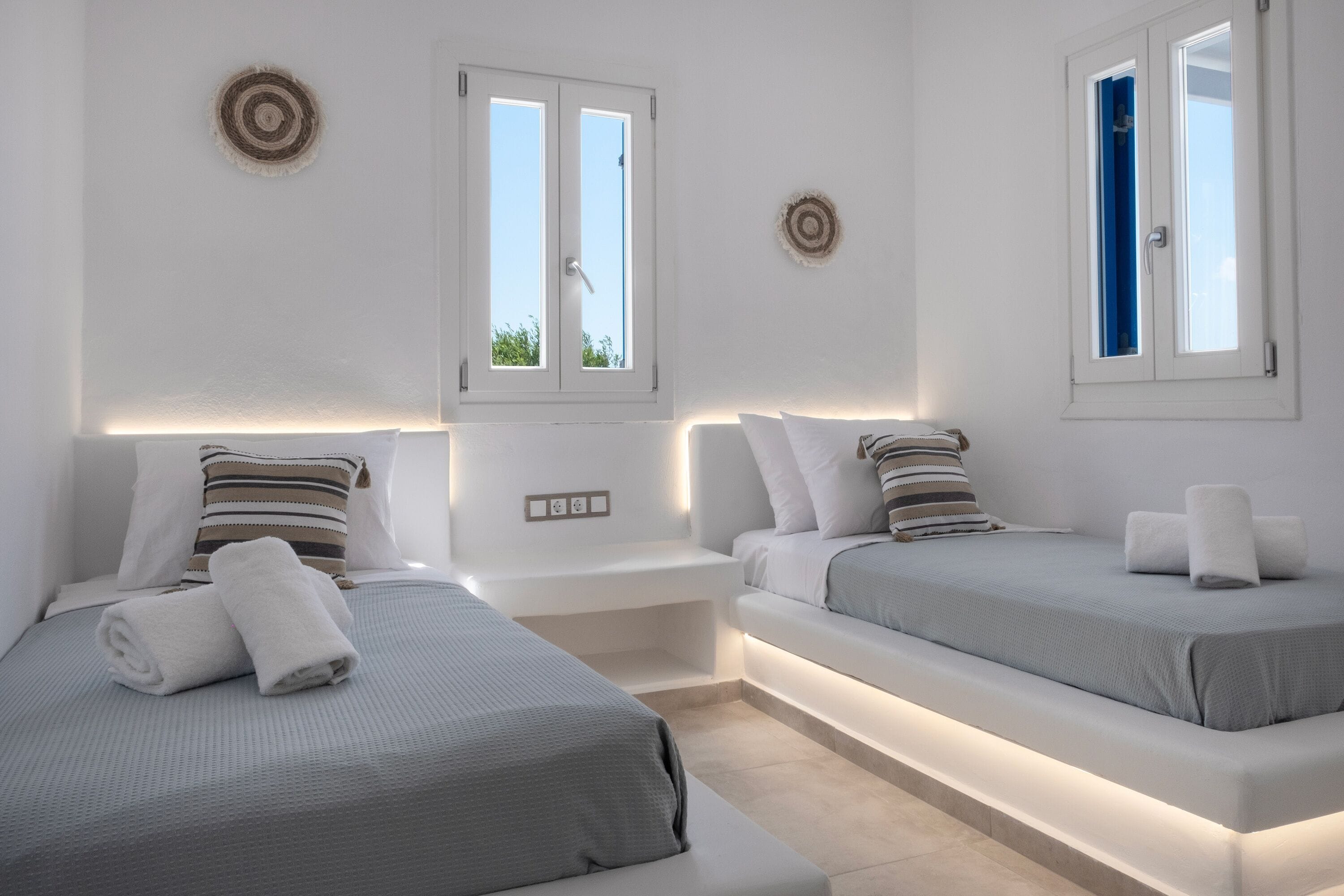 A modern hotel room with two beds, each with a pillow and a rolled towel. There are bedside tables with built-in lighting and a window with a view of the sky.