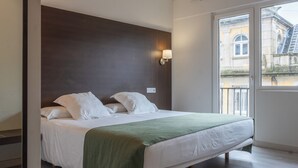 A modern bedroom with a large bed, a green throw blanket, a decorative ceiling light, and a view of a building through a large window. - Mendez Nunez (Lugo)