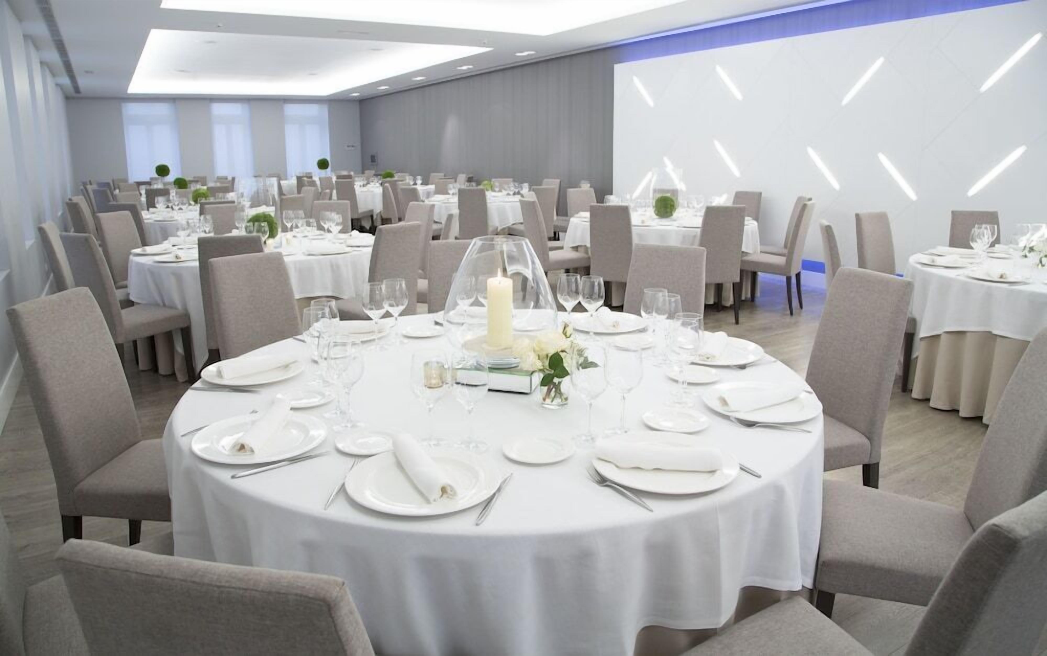 Un espai per a banquets amb taules rodones preparades per dinar, cadascuna amb mantells blancs, plats i coberts.