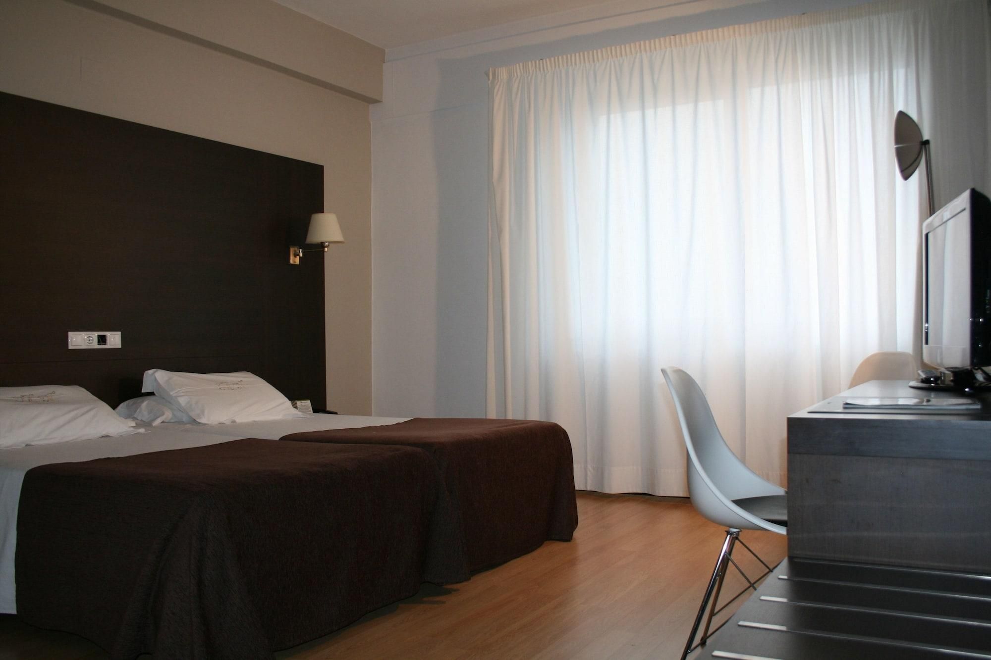 Una habitación de hotel con cama, dos mesitas de noche, un escritorio con silla, una lámpara y un televisor.