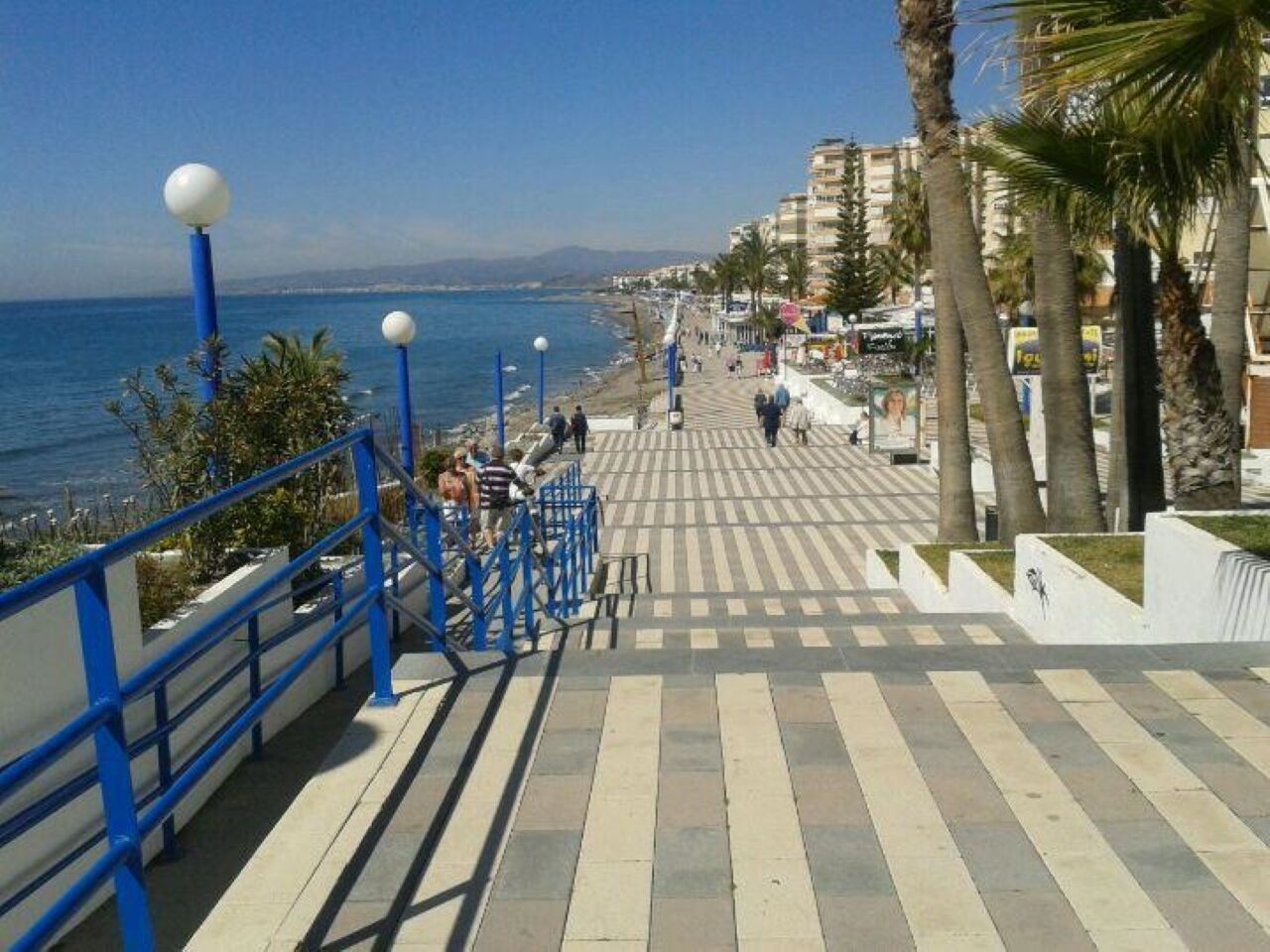 Un paseo marítimo con una escalera que baja a la playa, palmeras y una vista despejada del mar.