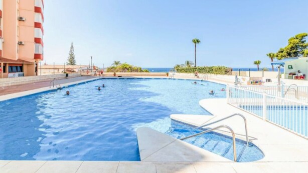 A swimming pool with multiple lanes, surrounded by a white fence, and a building with red and white stripes in the background. - Mango Estudio!! Piscina!! Playa a 150m!! (Torrox)