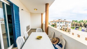 Un balcon avec une table et des chaises blanches, une porte à volets bleus et une vue sur des immeubles et des palmiers.