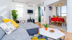 A modern living room with a grey sectional sofa, a flat-screen TV, and a dining area with a red tablecloth. - Sun City Apartment (Dubrovnik)