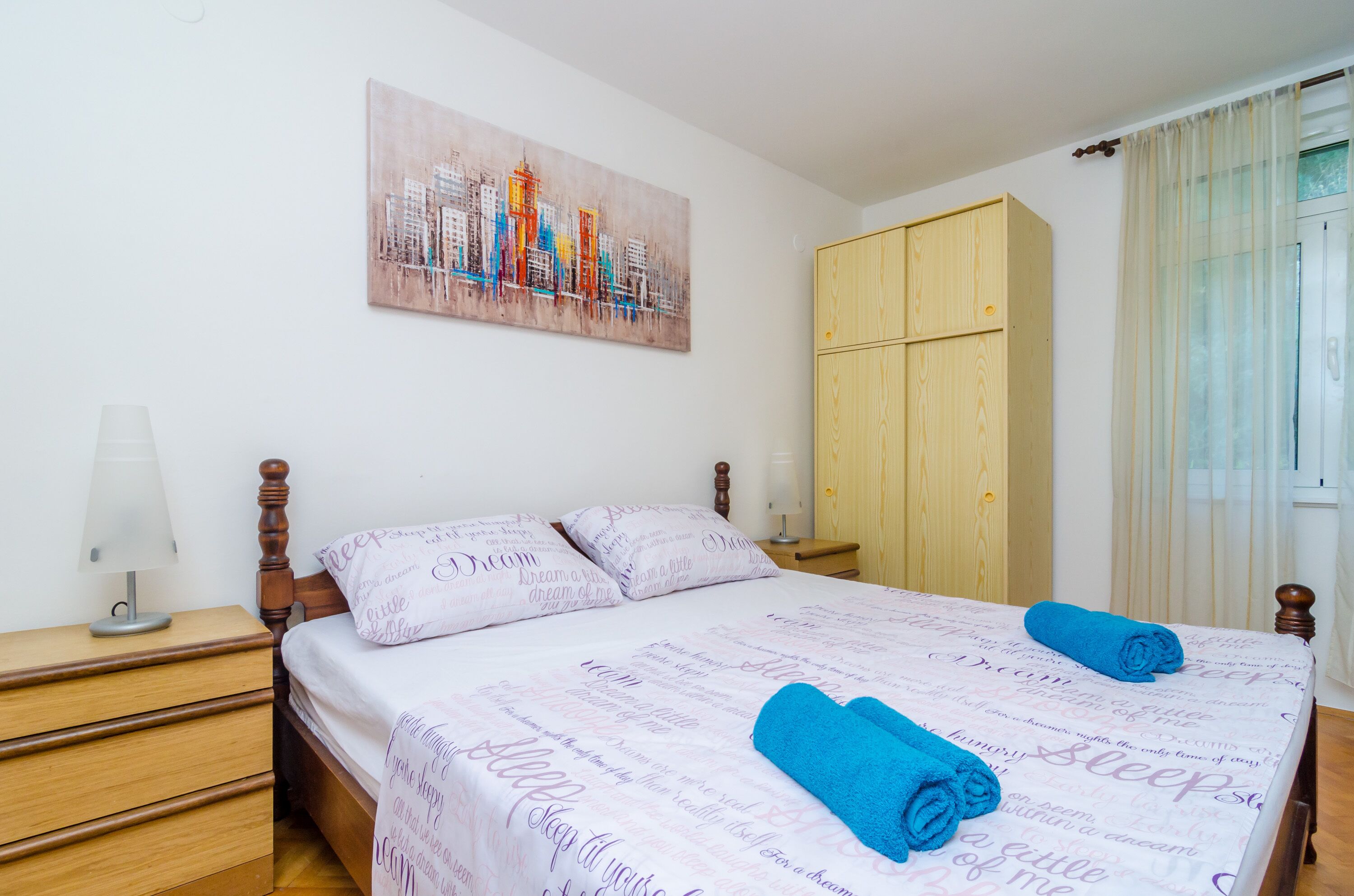A neatly made bed with a cityscape painting above it, a wooden wardrobe, and a nightstand with a lamp.