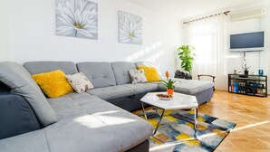 A modern living room with a grey sectional sofa, a white coffee table, and a colorful rug. There is a large window with sheer curtains, a flat-screen TV mounted on the wall, and a potted plant on a side table. - Sun City Apartment (Dubrovnik)