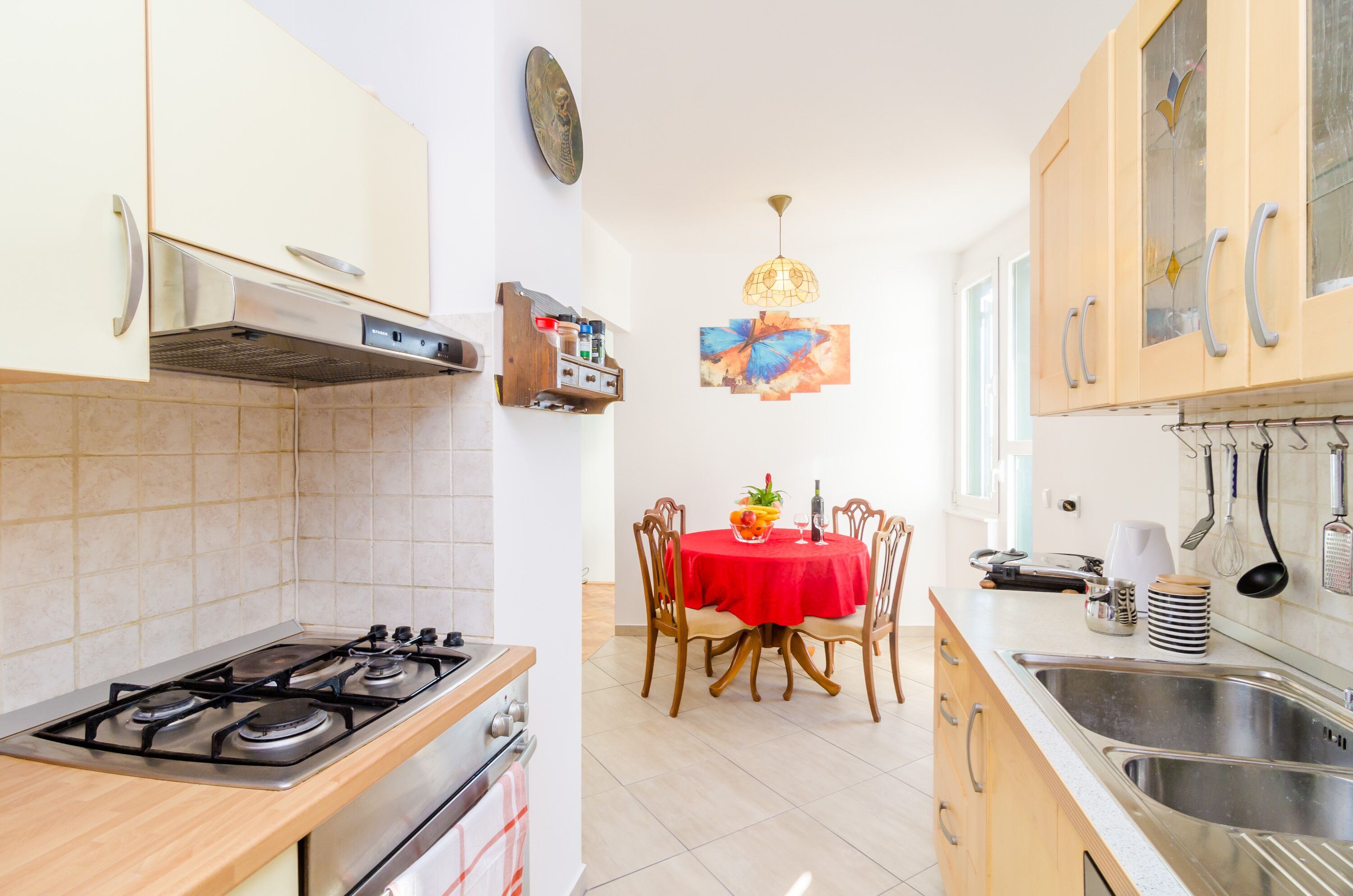A kitchen with a gas stove, oven, and sink. There is a dining table with chairs and a red tablecloth. A chandelier hangs from the ceiling.