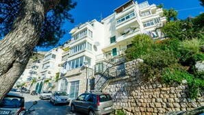 A row of white multi-story buildings with balconies, parked cars, and a stone wall. - Sun City Apartment (Dubrovnik)
