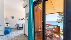 A balcony with a table and chairs, a view of the sea, and a door leading to an interior space with a desk and a bed. - Fantastic Seaview Turquoise House (Santa Margherita di Pula)