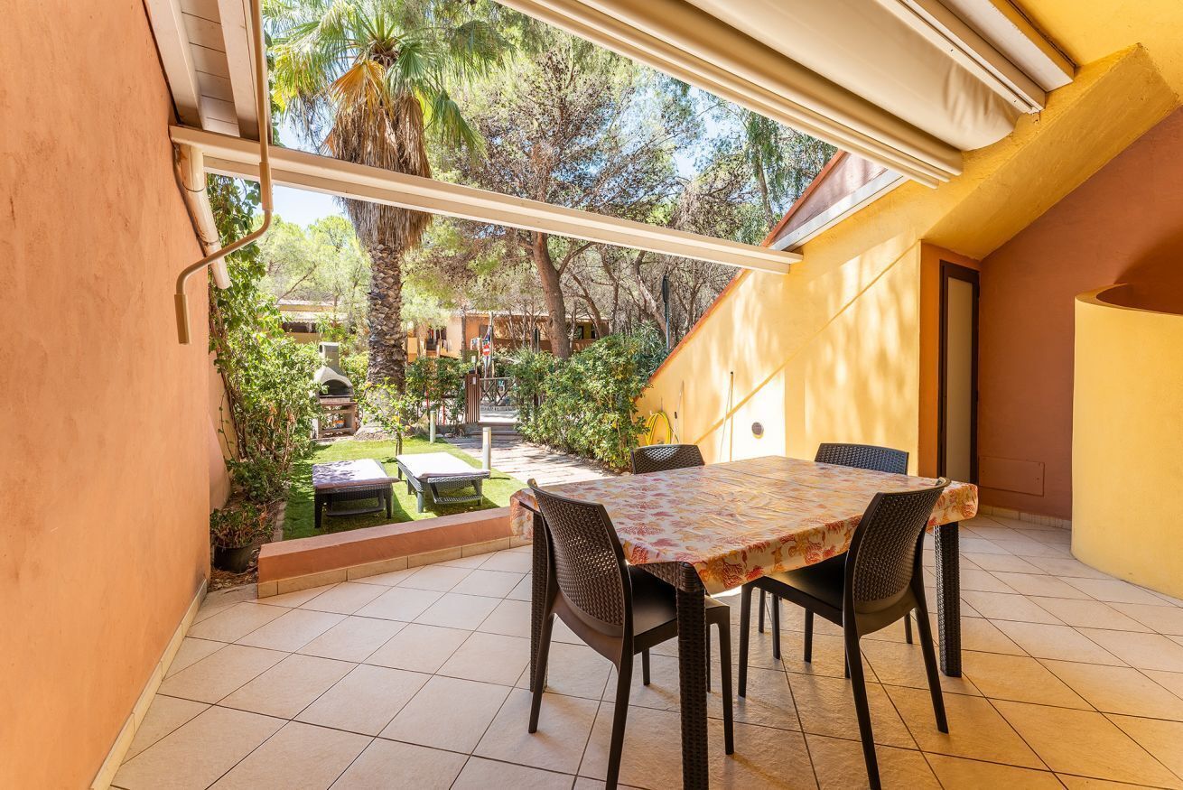 Um pátio com mesa e cadeiras, vista para um jardim e um edifício ao fundo.