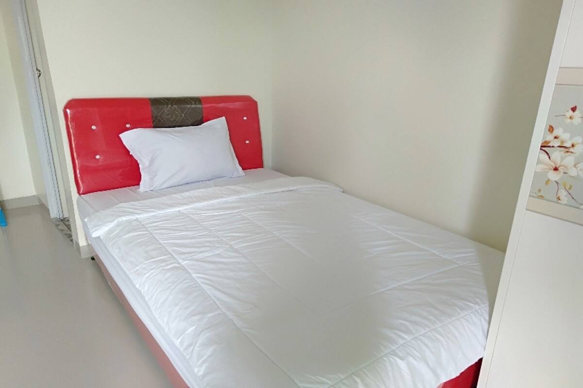 Ein Einzelbett mit rotem Kopfteil und weißer Bettwäsche in einem Raum mit schlichter weißer Wand und einer Tür links.