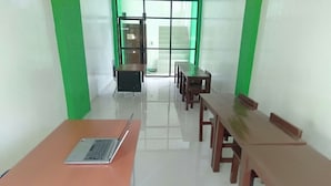 Ein Zimmer mit einem langen Tisch und Stühlen, einem Laptop auf dem Tisch und einem Fenster mit grünem Rahmen.
