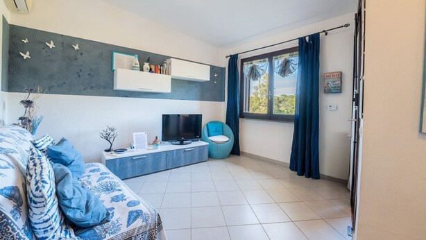 A modern living room with a sofa, TV, and a small table with a plant. There is a window with curtains and a view of greenery outside. - Blue House, 300 meters from the beach (Santa Margherita di Pula)