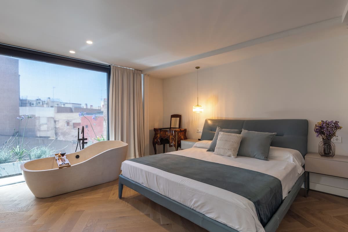 A modern bedroom with a large bed, a bathtub, and a view of buildings outside.
