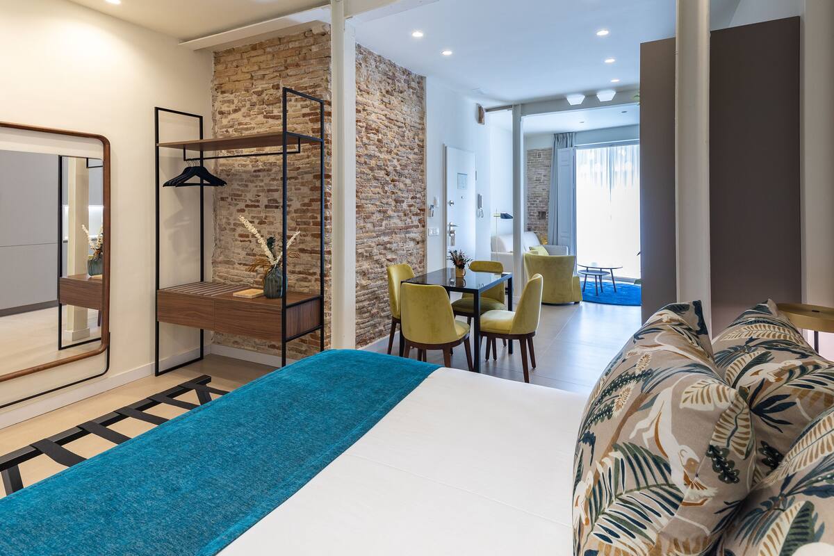 A modern hotel room with a large bed, a brick accent wall, a dining area with yellow chairs, and a view into the dining space.