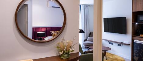 A modern hotel room with a round mirror, a wooden reception desk, a flat-screen TV, and a seating area with a yellow rug.
