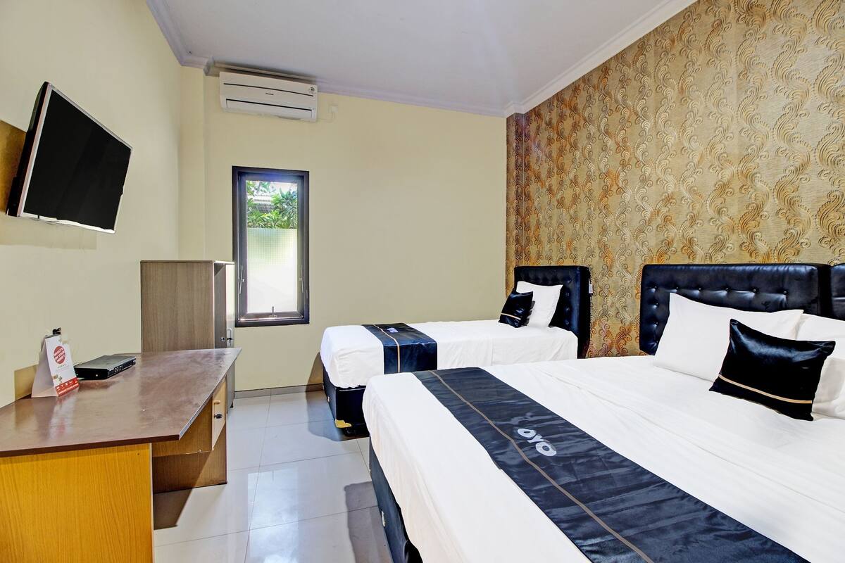 A hotel room with two beds, a desk, a flat-screen TV, and a window with a view of greenery.