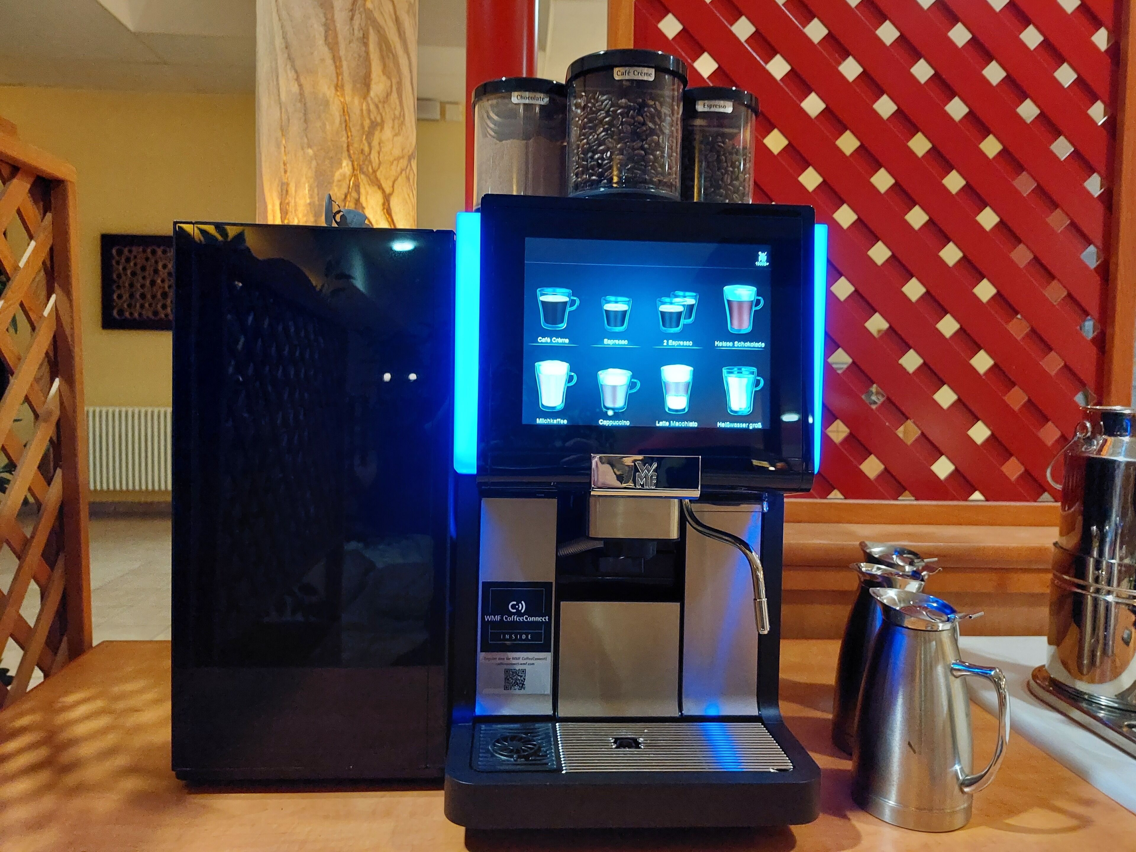 Una máquina de café con menú digital, una jarra de café y un espumador de leche sobre una mesa de madera.
