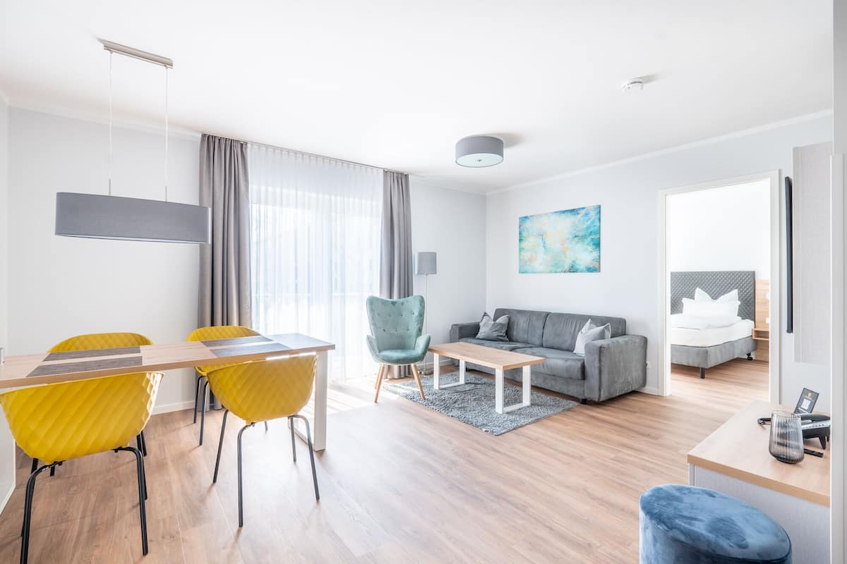 En moderne stue med spiseplads, en grå sofa, en blå lænestol og en seng med gråt sengegavl.