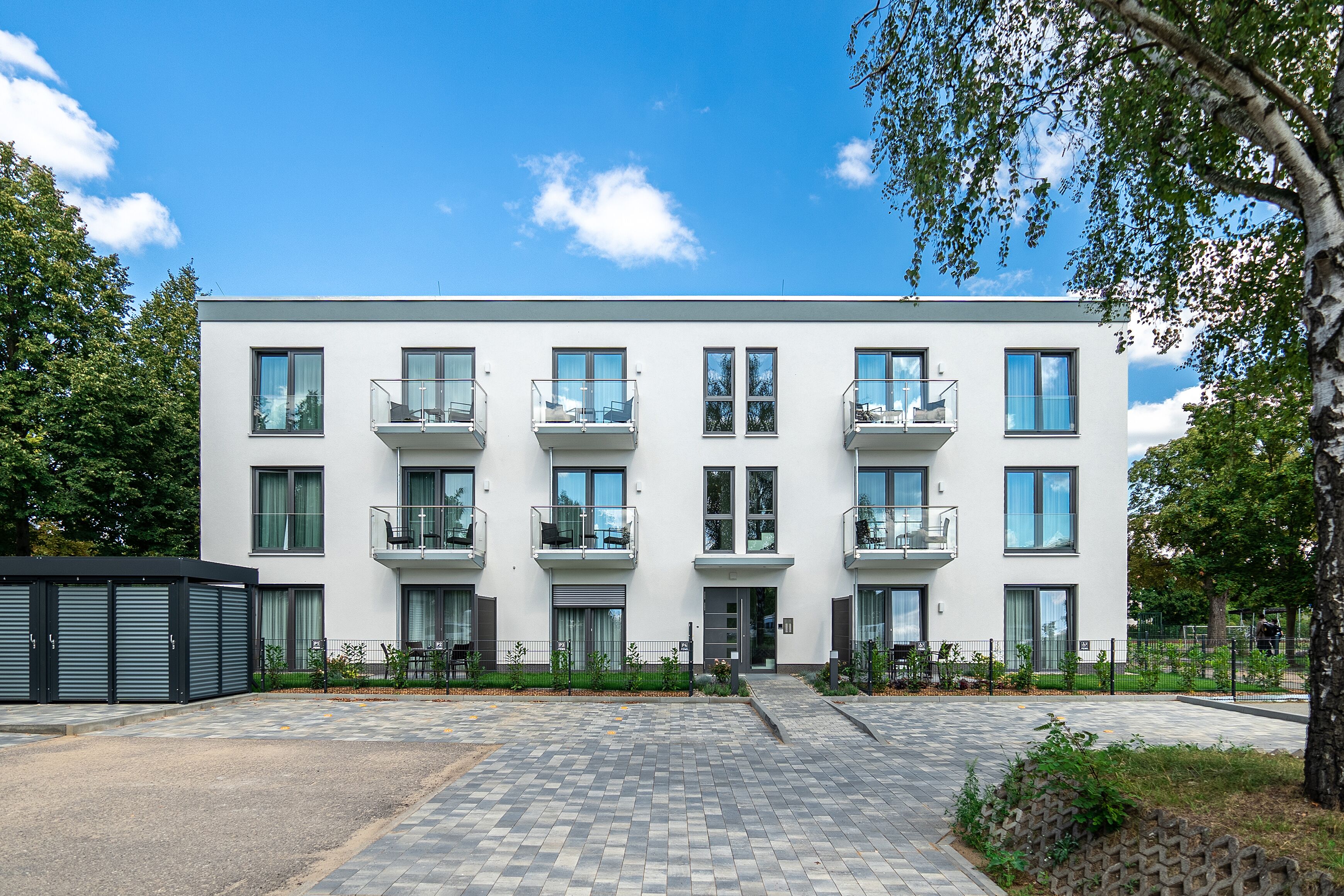 A modern multi-story building with balconies, a central entrance, and a paved courtyard.