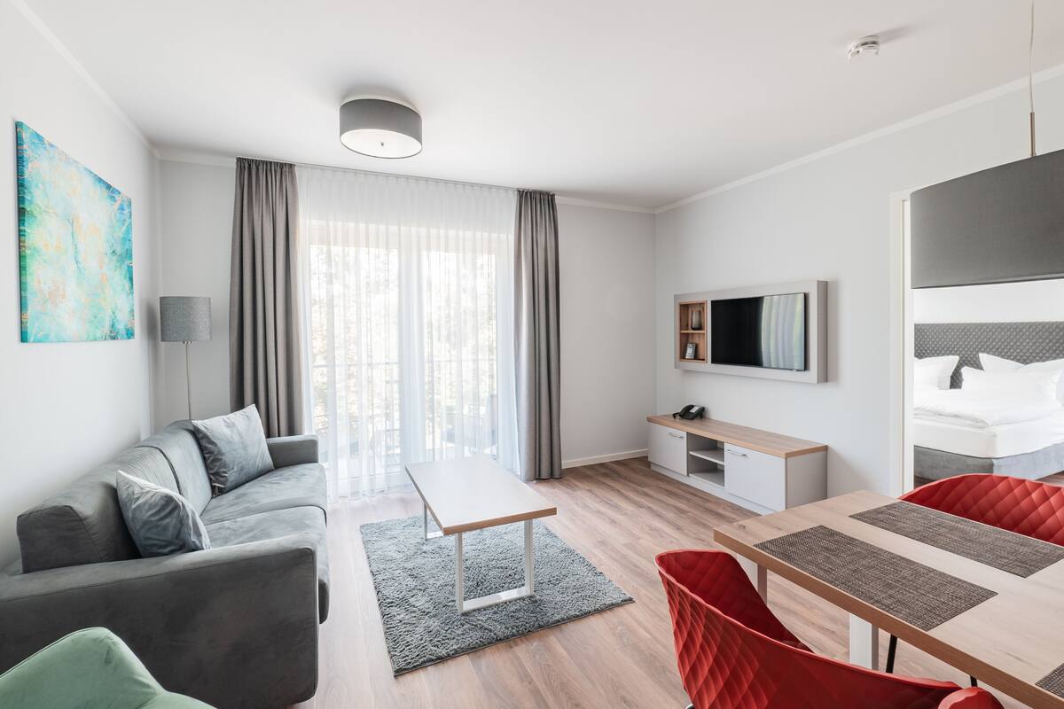 Et moderne hotelværelse med en grå sofa, et træspisebord, røde stole, et fjernsyn ophængt på væggen og et stort vindue med gardiner.