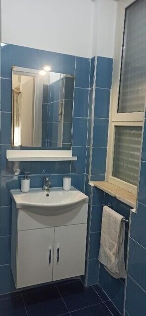 Une salle de bain avec un lavabo, un miroir et un meuble de rangement blancs, des carreaux bleus et une fenêtre avec des stores.