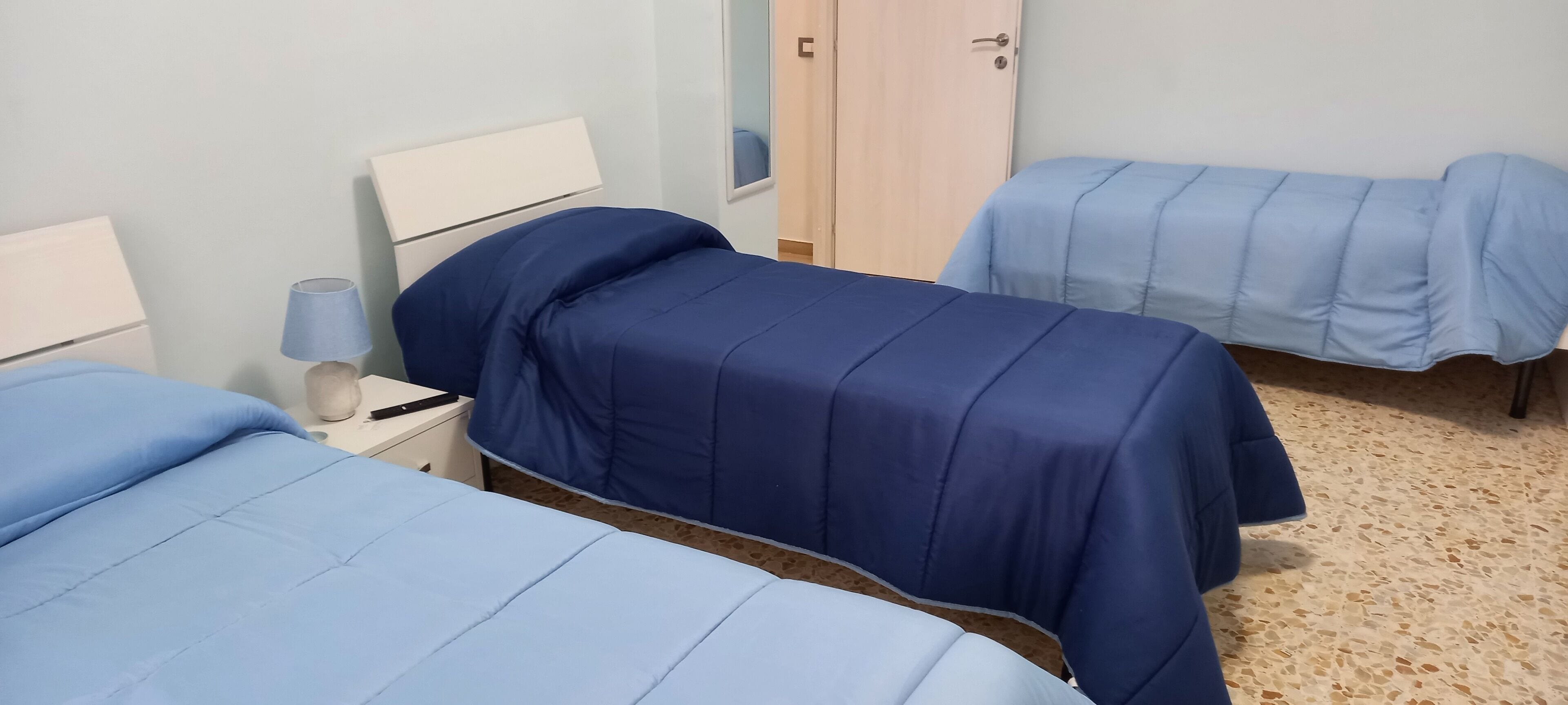 Ein Zimmer mit zwei Betten, jeweils mit blauer Bettdecke, einem Nachttisch mit Lampe und einem Holzschrank.