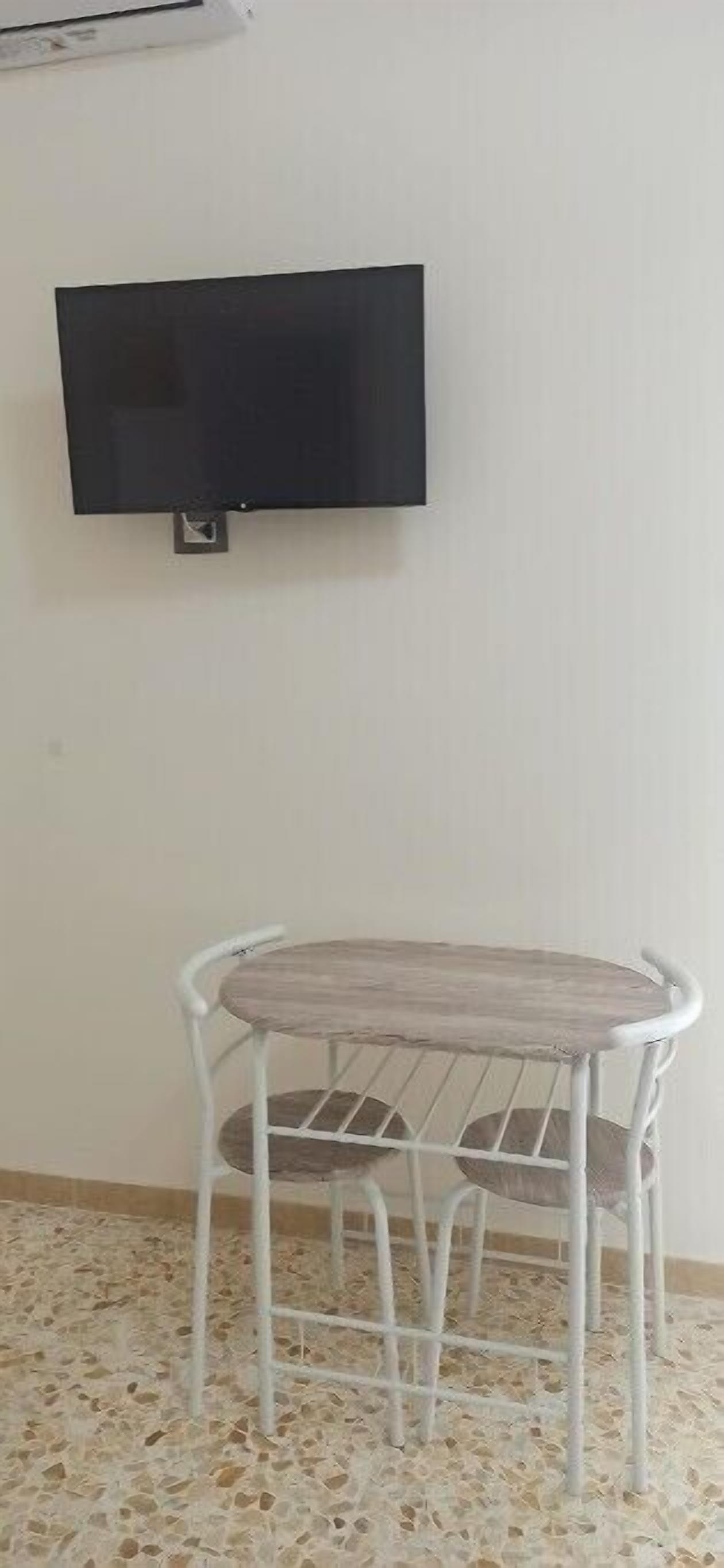Ein Zimmer mit weiß gefliestem Boden, einem runden Tisch mit Metallgestell und einem an der Wand montierten Flachbildfernseher.