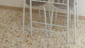 Una habitación con suelo de baldosas blancas, una mesa redonda con estructura metálica y un televisor de pantalla plana montado en la pared.