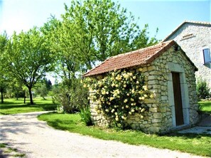 Edifici de pedra amb una entrada petita i un teulat de rajoles vermelles, envoltat de vegetació i arbres.