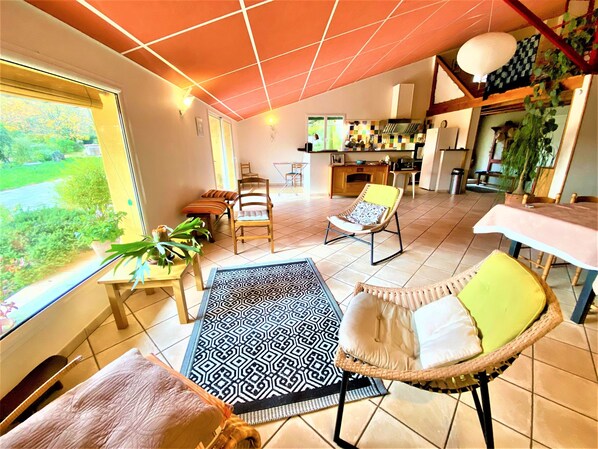 A spacious living area with a large window, a dining table, and a kitchen in the background.