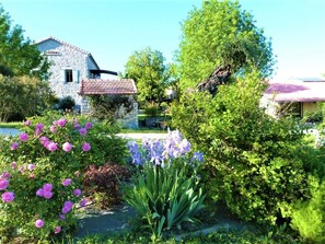 A stone house with a tiled roof, surrounded by lush greenery and flowering plants.