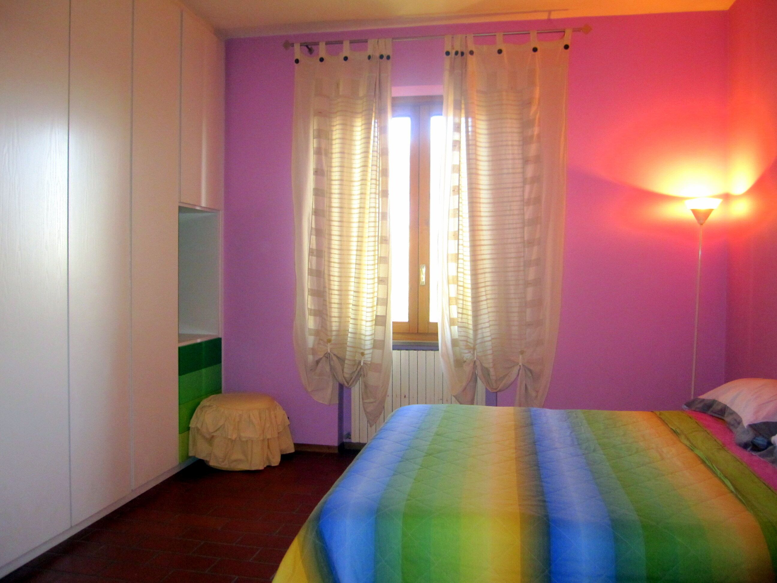 Ein Schlafzimmer mit Bett, einem Fenster mit Vorhängen, einer Nachttischlampe und einem kleinen Hocker.