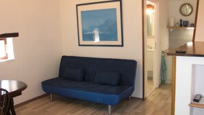 Ein Zimmer mit einem blauen Sofa, einem gerahmten Bild an der Wand, einem kleinen Tisch und einem durch eine offene Tür einsehbarer Küchenbereich.