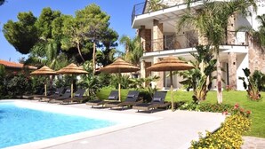 Zona de piscina con sillas de descanso y sombrillas, rodeada de vegetación y un edificio grande con un balcón.