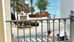 A balcony with a view of the sea, a street, and a building with palm trees. - Apartamentos Latin  (Nerja)