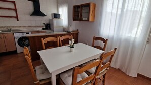 A compact kitchen with a dining area, including a table with chairs, a washing machine, a microwave, and a cabinet with glass doors. - Apartamentos Latin  (Nerja)