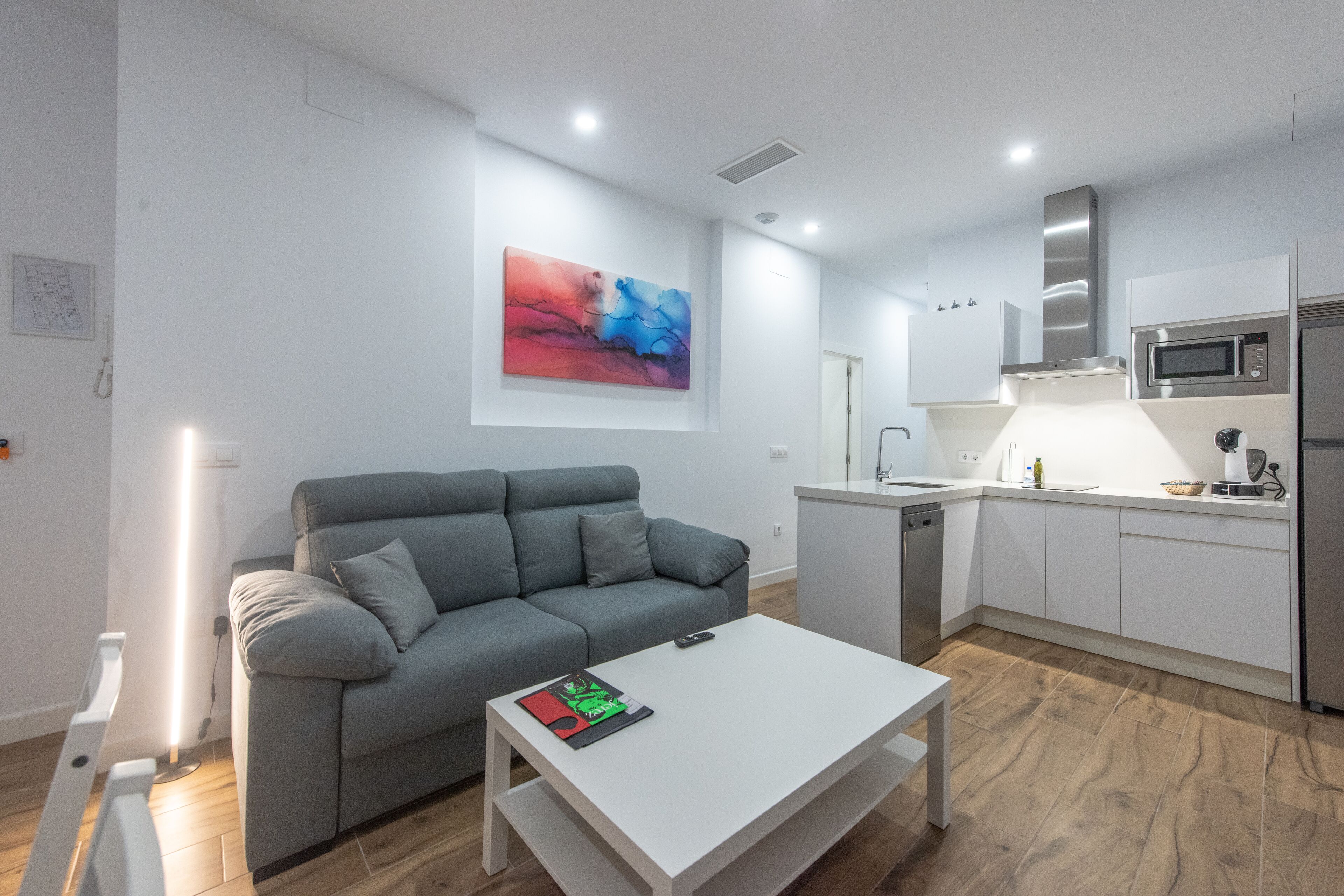 A modern living room with a grey sofa, a white coffee table, and a kitchen area with white cabinets and stainless steel appliances.
