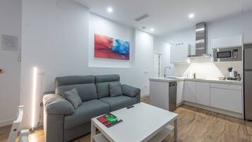 A modern living room with a grey sofa, a white coffee table, and a kitchen area with white cabinets and stainless steel appliances.