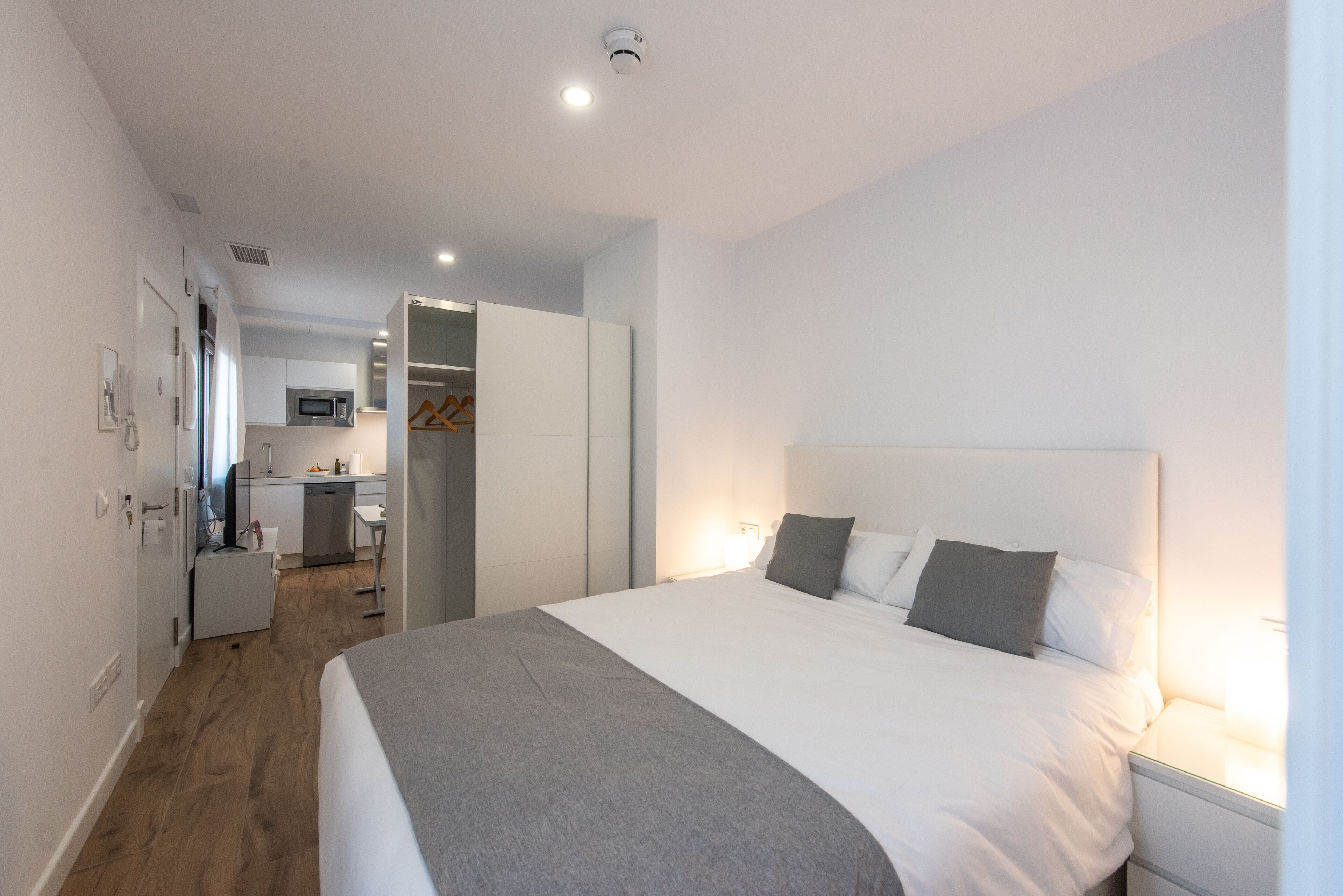 A hotel room with a large bed, a bedside table, a wardrobe, and a kitchenette in the background.