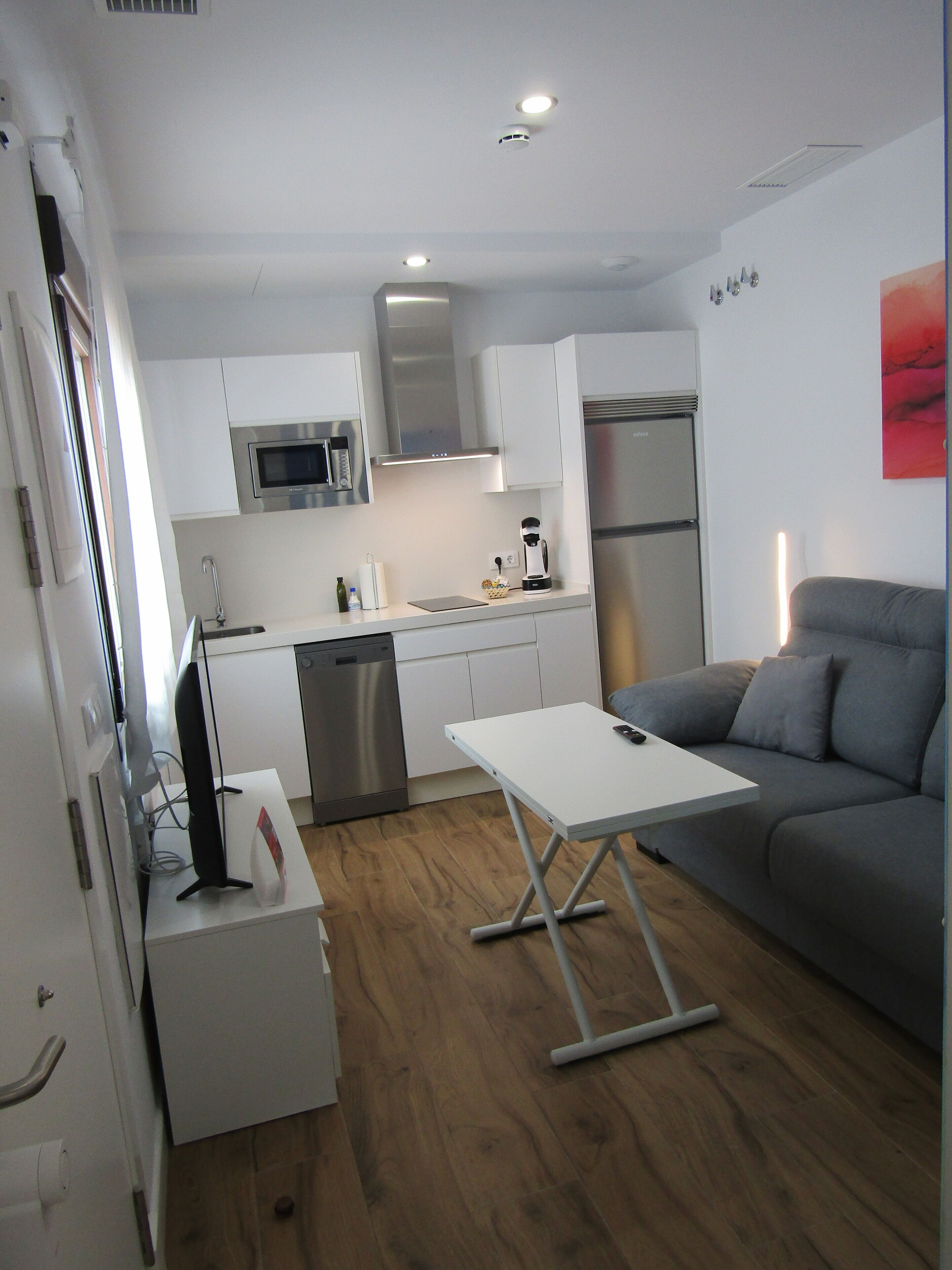 Un espacio habitable compacto con kitchenette, un sofá gris y una pequeña mesa.