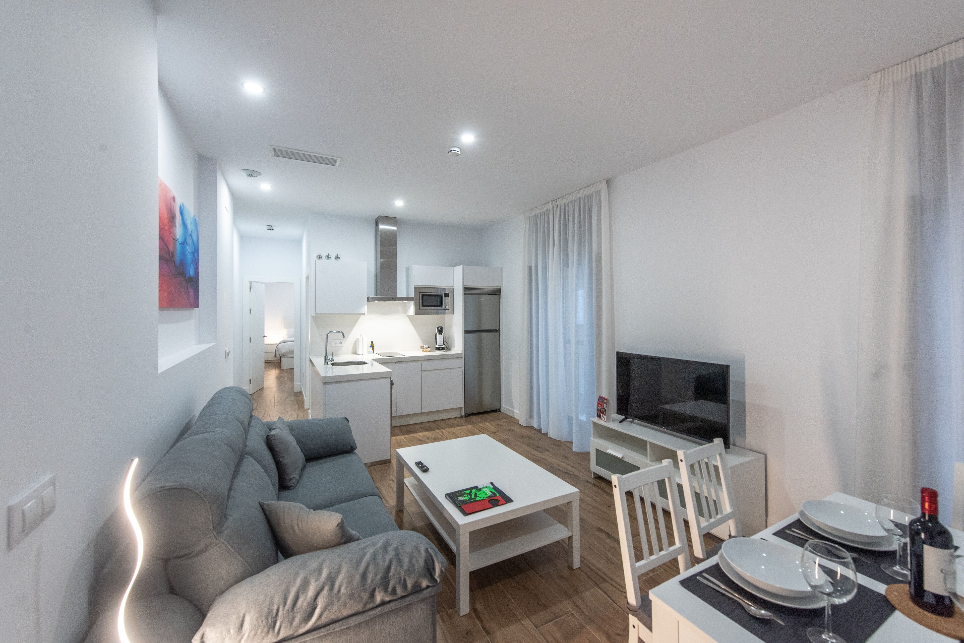 A modern living room with a grey sofa, a white coffee table, and a TV. There is a kitchen area with a microwave and a refrigerator.