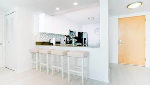 Une cuisine moderne avec des meubles blancs, un pare-feu en carrelage et un four encastré. On y trouve trois tabourets de bar blancs et une porte en bois.